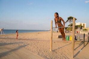 Ein Strand mit einer Klimmzugstange und Trainingsgeräten, eine Person macht Klimmzüge, und eine weitere Person steht im Sand.