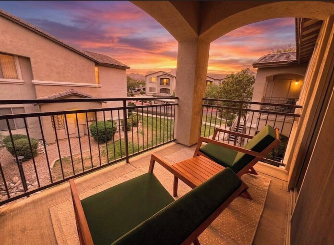 Un balcone con due poltrone imbottite verdi e un tavolo di legno, con vista su una zona residenziale con case e un tramonto.