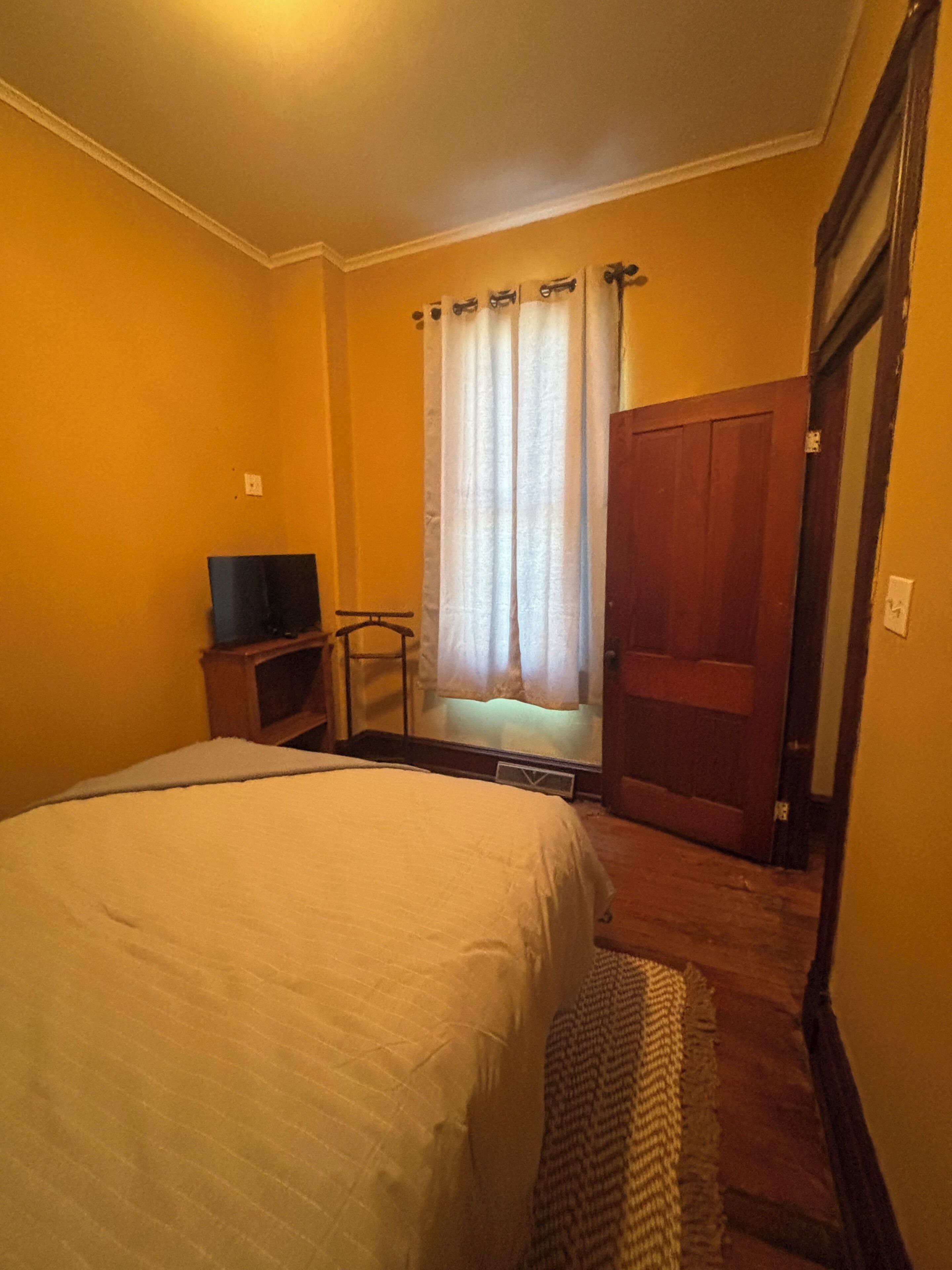 A bedroom with a bed, a television on a stand, a window with curtains, and a door.