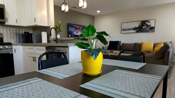 Une cuisine moderne avec des meubles blancs, une table à manger avec des sets de table, un vase jaune avec une plante, et un coin salon avec un canapé et des coussins.