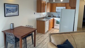 A compact kitchen with wooden cabinets, a microwave, a stove, and a refrigerator. There is a dining table with chairs and a sofa in the foreground. - Charming 1-bedroom condo nestled in the mountains of Heavenly Ski Resort (Stateline)