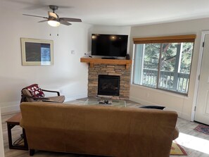 A living room with a stone fireplace, a flat-screen TV mounted above it, a brown sofa, and a ceiling fan. - Charming 1-bedroom condo nestled in the mountains of Heavenly Ski Resort (Stateline)