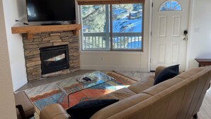 A living room with a stone fireplace, a flat-screen TV mounted on a wooden shelf, a glass coffee table, a beige sofa, and a view of a snowy landscape through a window. - Charming 1-bedroom condo nestled in the mountains of Heavenly Ski Resort (Stateline)