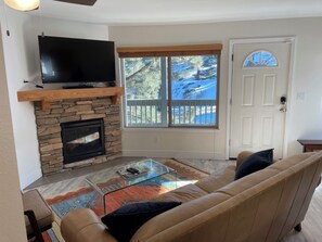 Ein Wohnzimmer mit Kamin aus Stein, einem an einer Holztrennwand montierten Flachbildfernseher, einem Glastisch, einem beigefarbenen Sofa und Blick auf eine verschneite Landschaft durch ein Fenster.