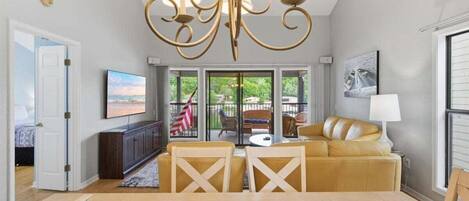 A spacious living room with a large dining table, yellow sofas, a chandelier, and a television.