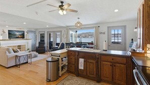 A spacious living room with a fireplace, a dining area, a kitchen with wooden cabinets, and a large window offering a view of the outdoors. - Zero-step Dock Life With Big Views, Lake Toys, Firepit, Boat Slip at 43mm (Roach)