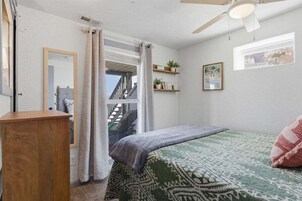 Un dormitorio con cama, cómoda de madera, ventilador de techo, ventana con cortinas y una pequeña estantería con plantas.