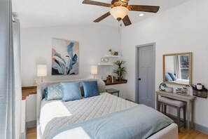 Ein Schlafzimmer mit Bett, Deckenventilator, Gemälde, Nachttisch, Spiegel und Tür.