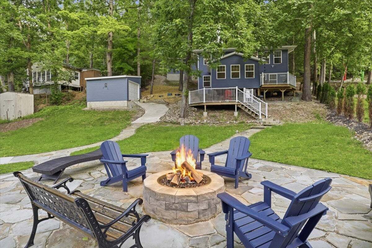 Un patio de piedra con un hogar a leña y sillas Adirondack, rodeado de un césped cuidado y una casa con terraza.