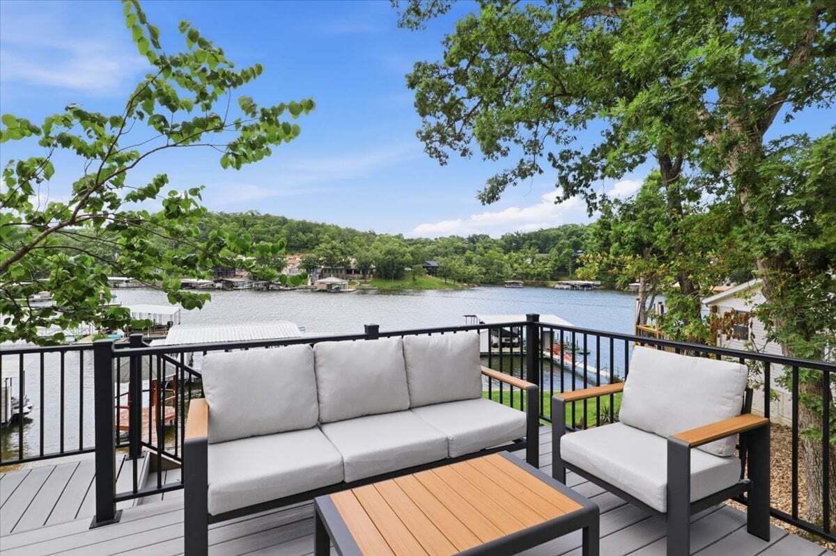 Terraza con sofá y sillones, con vista a un lago y embarcaciones, con casas al fondo.