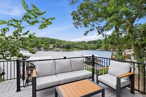 A deck with a sofa and chairs, overlooking a lake with boats and houses in the background.