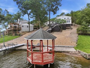 Um gazebo com telhado, um pequeno cais e uma casa ao fundo.