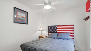 Un dormitorio con cama, ventilador de techo, mesita de noche, lámpara y cabecera con la bandera estadounidense.