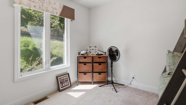 Una habitación con un ventanal grande, un mueble de almacenamiento de mimbre, un ventilador de pie y una cita inspiradora enmarcada en el suelo.