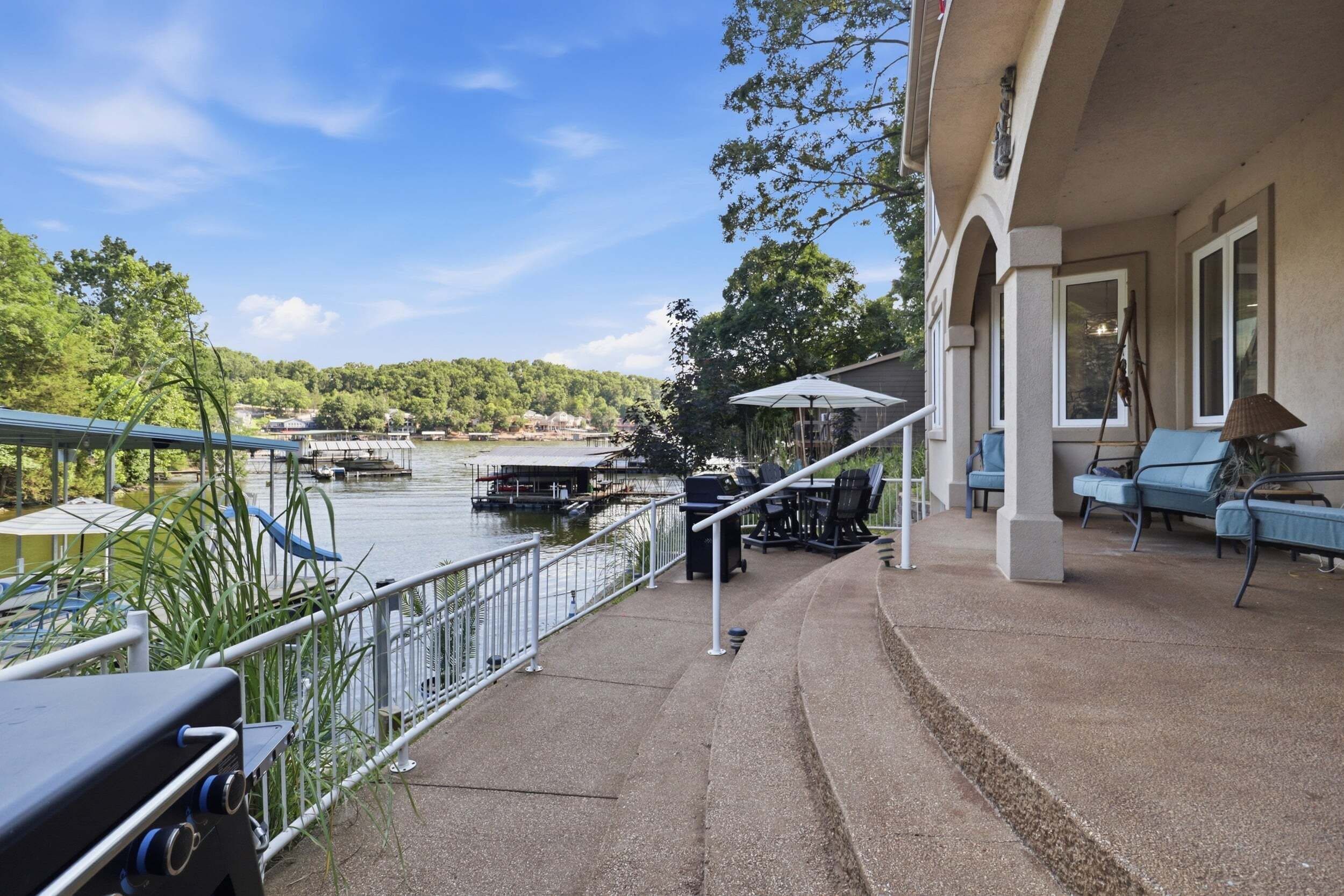 A waterfront patio with outdoor furniture, a grill, and a view of the lake and boats.
