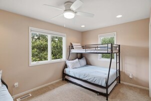 Habitación con literas, ventilador de techo, dos ventanas con vistas a árboles y piso alfombrado.