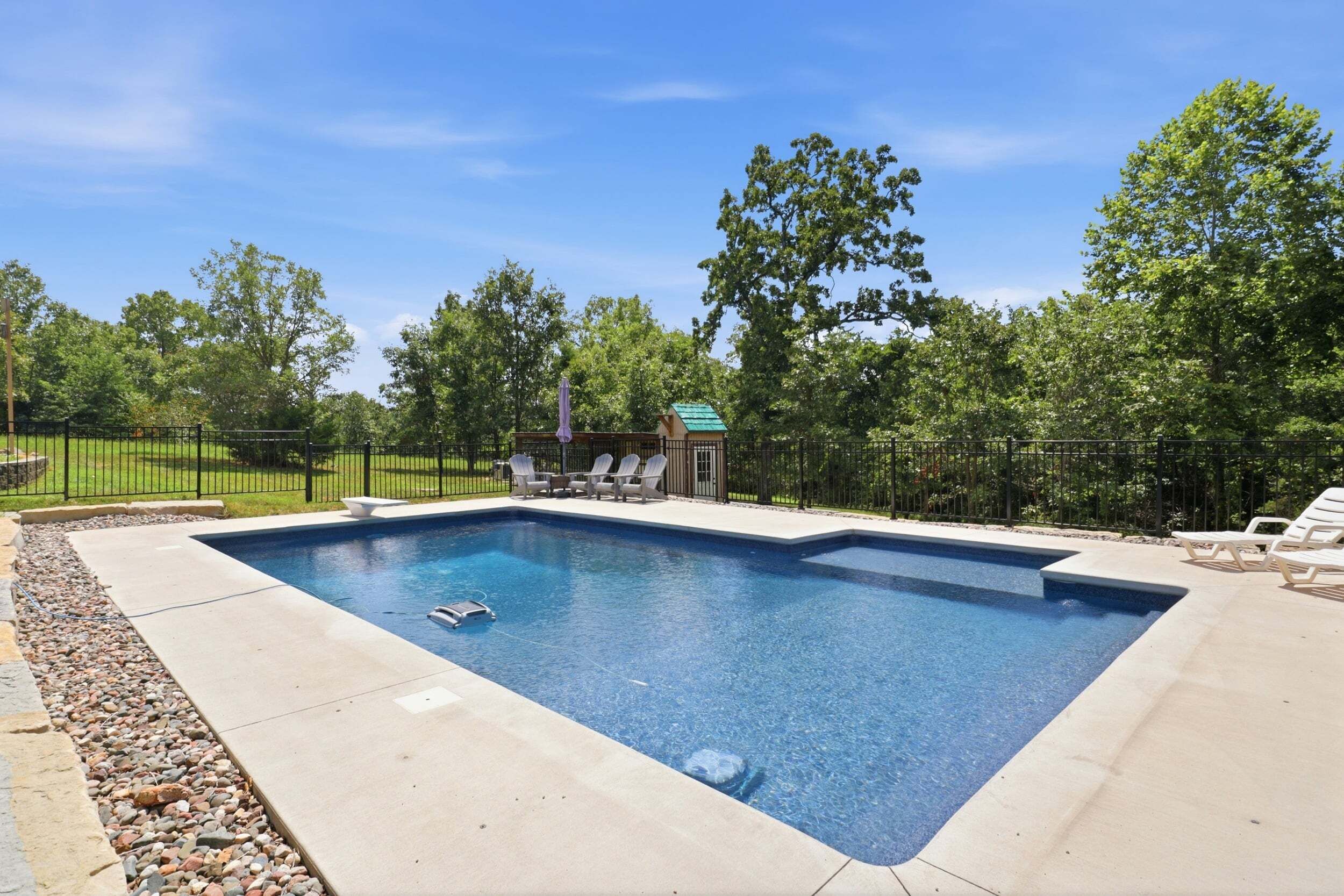 A rectangular swimming pool with a built-in hot tub, surrounded by a deck with lounge chairs, a small covered seating area, and a fence.