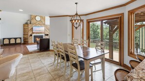 A dining area with a wooden table and chairs, a fireplace, and a large window with a view of greenery. - Private Heated Pool, Sleeps 18, Game Room & Outdoor Living (Camdenton)