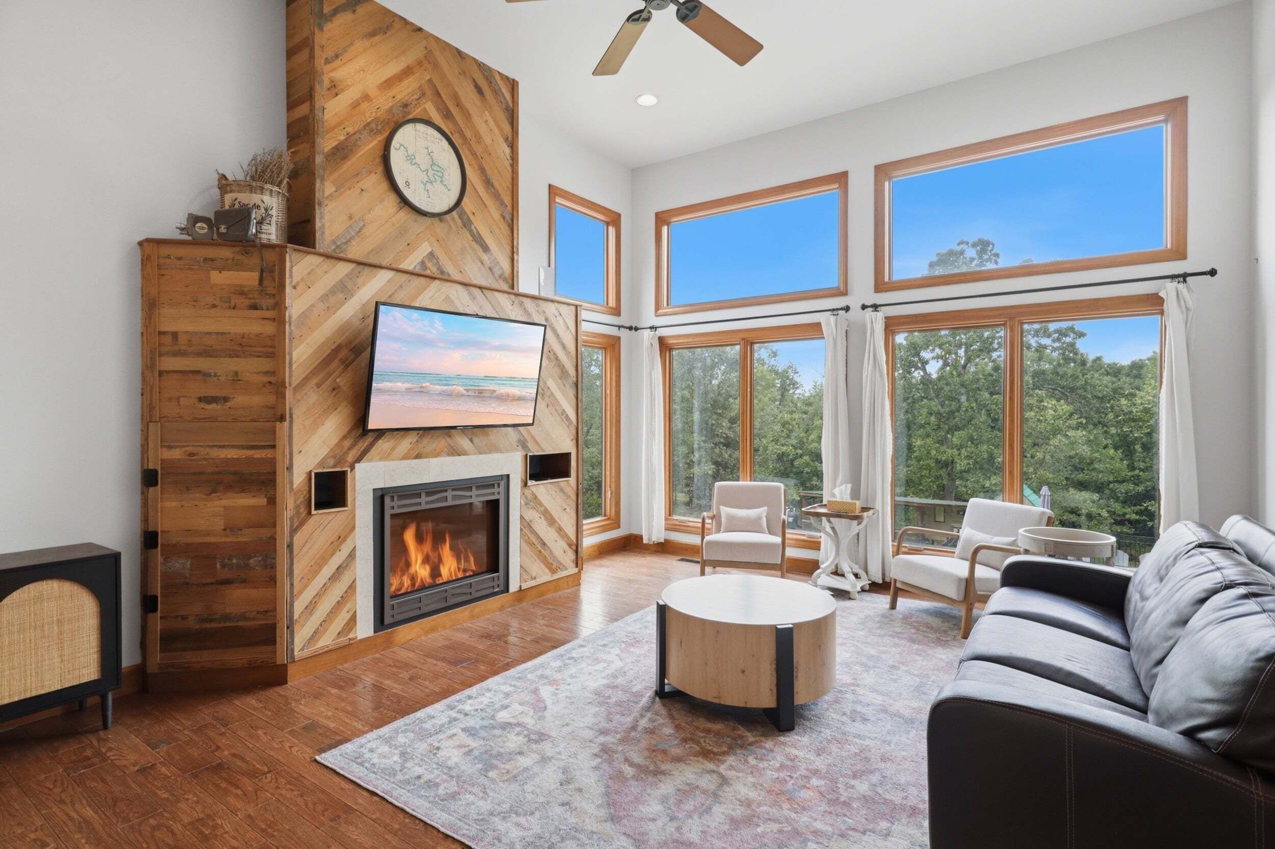 A spacious living room with a fireplace, large windows, and a ceiling fan.