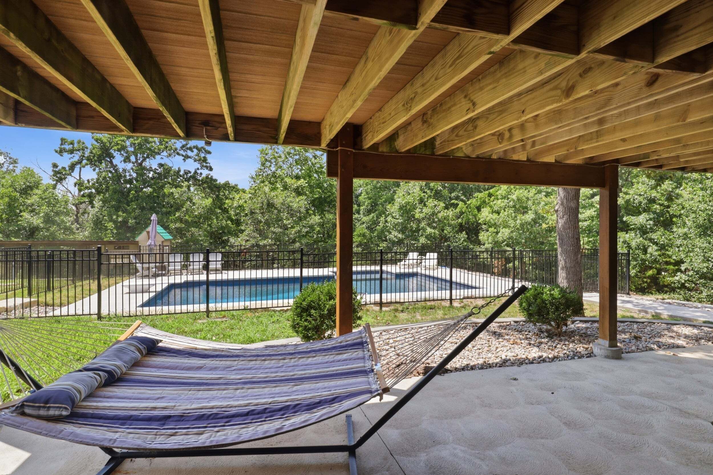 A hammock under a wooden pergola with a pool and a fenced yard in the background.