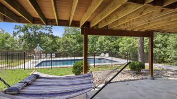 A hammock under a wooden pergola with a pool and a fenced yard in the background.