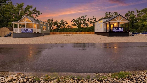 Two small cabins with 'RENT ME!' signs, a gravel area, and a body of water in the foreground.