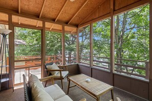 Un espacio de estar al aire libre cubierto con una mesa y sillas de madera, y ventanas amplias que dan a un paisaje de árboles.