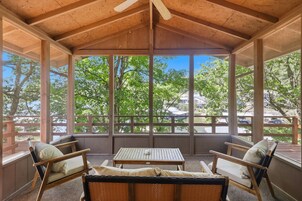 Un área de estar al aire libre techada con una mesa, sillas, ventanas amplias y vistas a árboles y edificios.