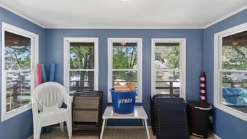 Una habitación con tres ventanas, una silla blanca, una mesita pequeña y un cubo de basura azul con la etiqueta 'JENGA'.