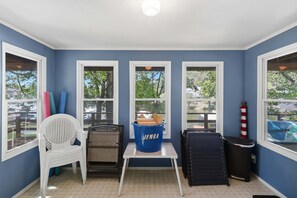 Una habitación con tres ventanas, una silla blanca, una mesita pequeña y un cubo de basura azul con la etiqueta 'JENGA'.