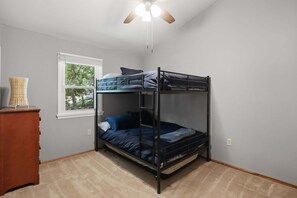 Een slaapkamer met stapelbed, plafondventilator, raam met zonwering en een houten dressoir.