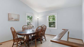 Un comedor con una mesa y sillas de madera, dos ventanas con marcos blancos y un cuadro en la pared.