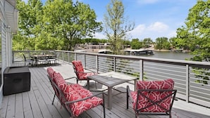 A deck with outdoor seating, a view of a river, and a building in the background. - Big Lakefront Lodge With Private Dock, 2 Slips, Game Room, Firepit & More at 11mm (Sunrise Beach)