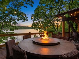 Uma mesa redonda ao ar livre com lareira embutida, rodeada por cadeiras de vime, sob uma pérgula com luzes de cordão, com vista para um lago.