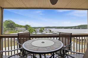 Uma varanda com uma mesa redonda e cadeiras, com vista para um lago e edifícios ao longe.