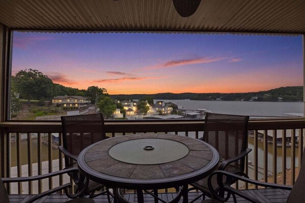 Uma varanda com uma mesa redonda e cadeiras, com vista para um lago e edifícios ao fundo.