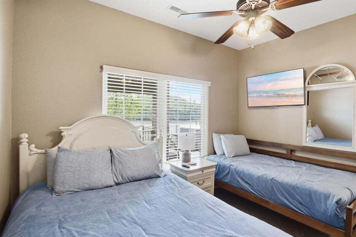 A bedroom with two beds, a ceiling fan, a window with blinds, a bedside table with a lamp, and a painting on the wall.