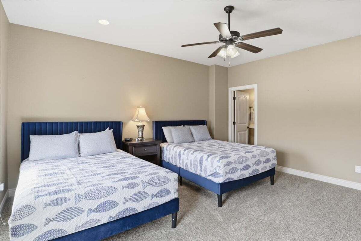 A bedroom with two beds, a ceiling fan, a nightstand with a lamp, and a door leading to another room.