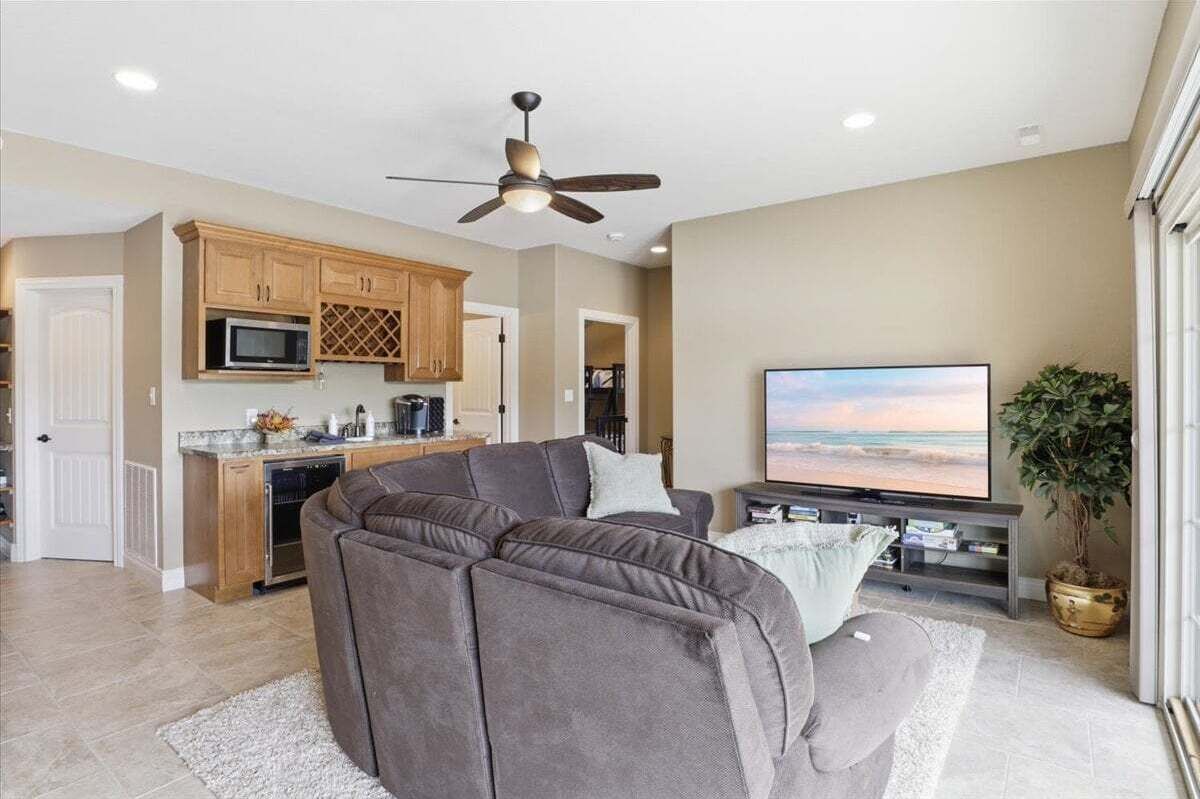 A living room with a sectional sofa, a flat-screen TV, a ceiling fan, and a kitchen area with wooden cabinets and a microwave.