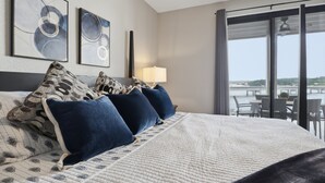 A hotel room with a large bed, two wall-mounted artworks, a nightstand with a lamp, and a view of the outdoors through a sliding door. - Main Channel Views, Resort Pools & Boat Slip at Topsider's 19mm (Osage Beach)