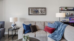 A living room with a grey sofa, a side table with a lamp, and a wall-mounted sign with the letters 'LKE'. - Main Channel Views, Resort Pools & Boat Slip at Topsider's 19mm (Osage Beach)