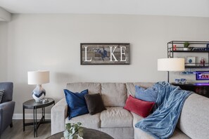 Ein Wohnzimmer mit einem grauen Sofa, einem Beistelltisch mit Lampe und einem an der Wand befestigten Schild mit den Buchstaben 'LKE'.