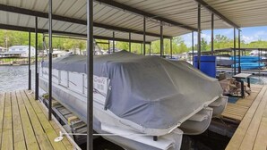 A covered dock with a boat covered by a tarp, a wooden deck, and a view of a water body with houses in the background. - Lakefront Fun With Private Dock, Firepit, Swim Mat, Kayaks & Screened Lake Room (Gravois Mills)