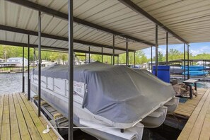 A covered dock with a boat covered by a tarp, a wooden deck, and a view of a water body with houses in the background. - Lakefront Fun With Private Dock, Firepit, Swim Mat, Kayaks & Screened Lake Room (Gravois Mills)
