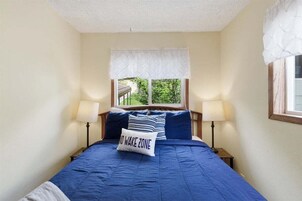 Un dormitorio con cama, dos lámparas de noche, una ventana con vista a follaje y un almohadón con la inscripción 'NO WAKE ZONE'.