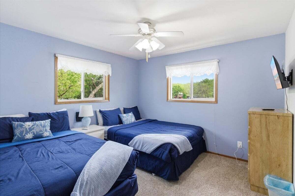 Un dormitorio con dos camas, un ventilador de techo, un armario de madera y una ventana con vistas a árboles.