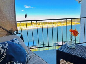 A balcony with a view of a beach and the sea, a sofa with patterned cushions, and a small table with a vase of flowers. - Right on the water! Lake Travis (Spicewood)