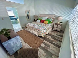 Ein Schlafzimmer mit Bett, Nachttischen, Überbett, einer Zimmerpflanze, einem Teppich und einem Fenster mit Jalousien.