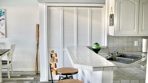 Una cocina con encimera blanca, un lavamanos y un florero verde. Hay sillas de madera y una mesa.
