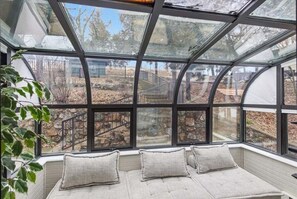 Ein Zimmer mit einem großen Glasfenster, einer Bank mit Kissen und Blick auf einen Kiesweg und Bäume.